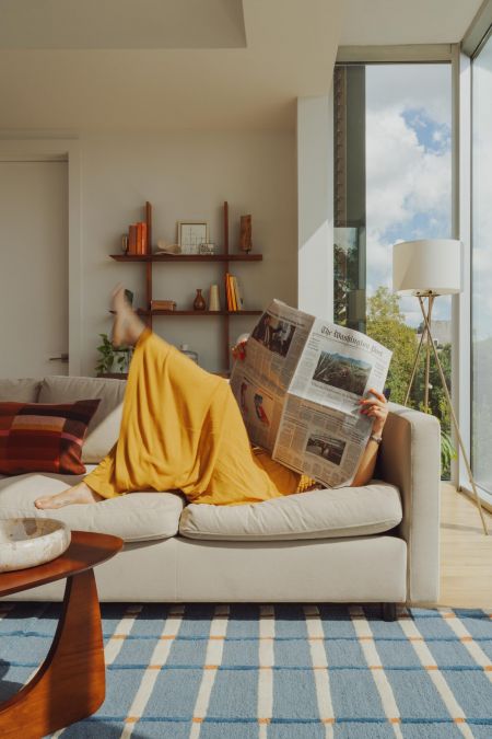 OSLO Coliving person in a yellow dress lounges on a sofa reading a newspaper in a bright, modern living room.