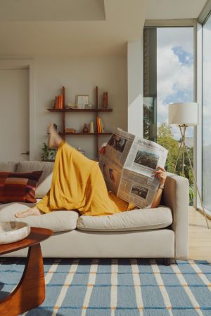 OSLO Coliving person in a yellow dress lounges on a sofa reading a newspaper in a bright, modern living room.